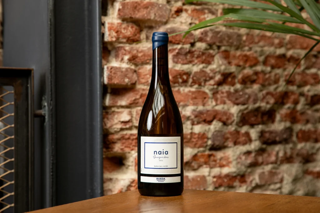 Botella de vino Sauvignon Blanc de Naia sobre una mesa de madera con una paed de ladrillos de fondo. Esta imagen se utiliza para ilustrar una entrada sobre qué es el vino sauvignon blanc.