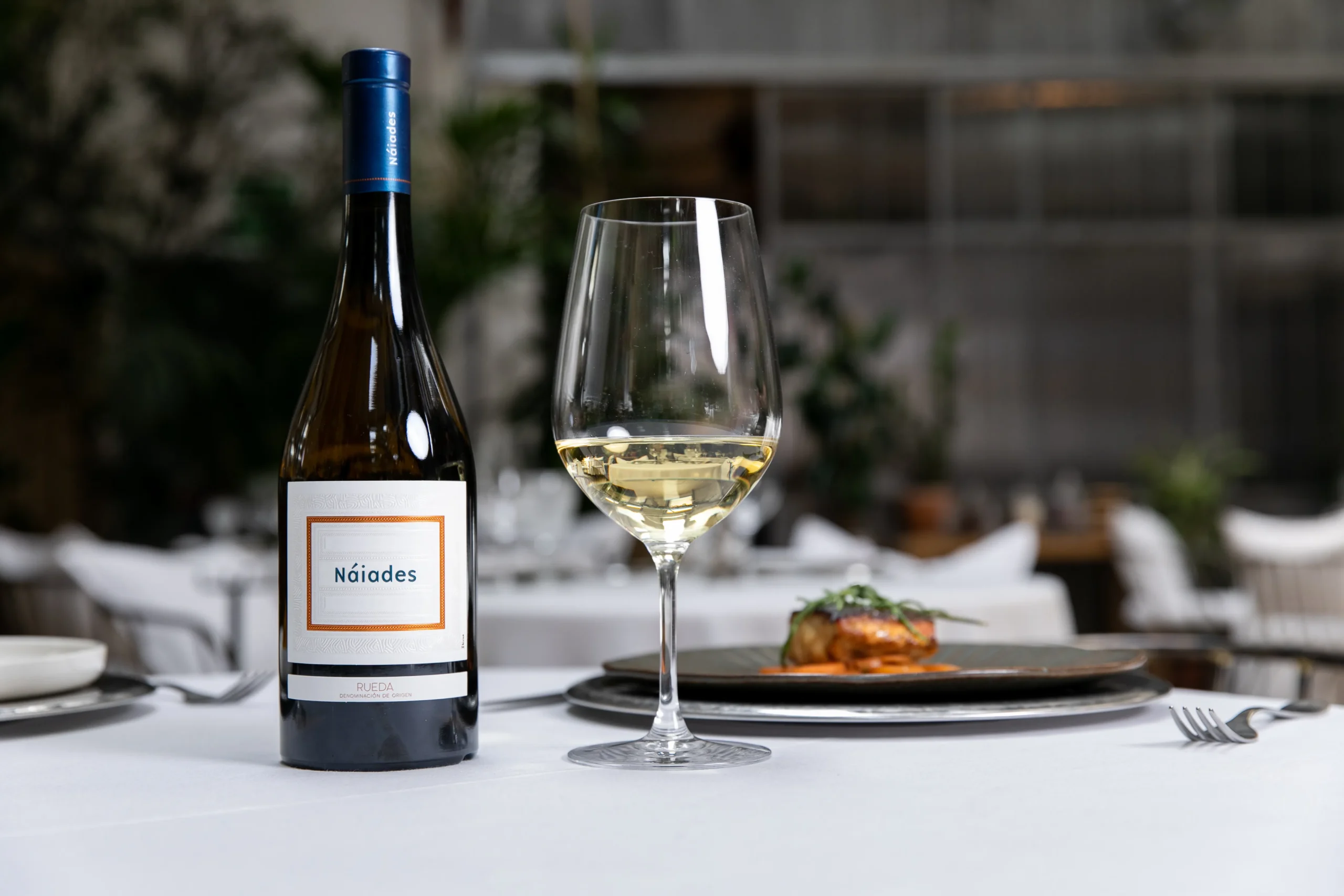 Botella de vino Náiades junto a una copa que contiene vino y un plato con una opción de maridaje.