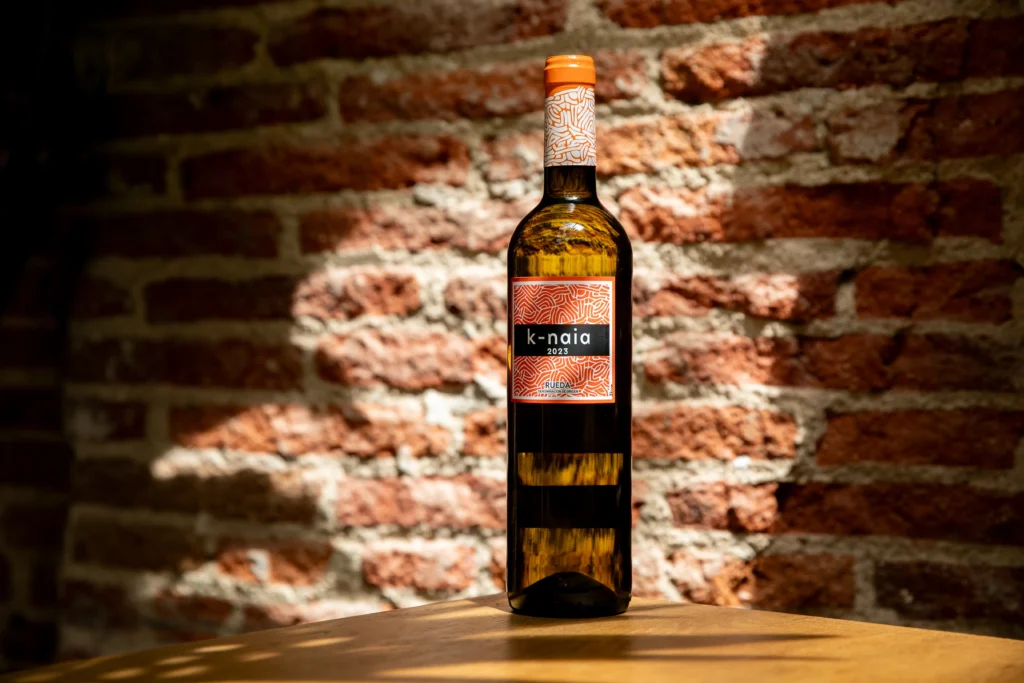 Botella de K-Naia situada sobre una mesa de madera con un fondo de una pared de ladrillo.